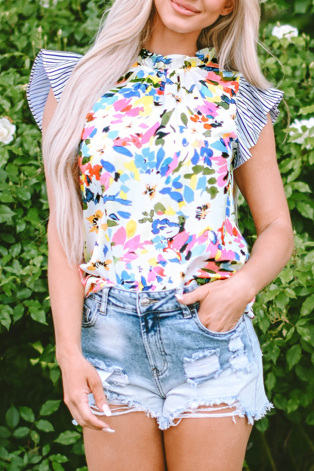 A woman wearing a light blue floral print blouse with striped ruffled sleeves and denim shorts, standing in front of a greenery background.