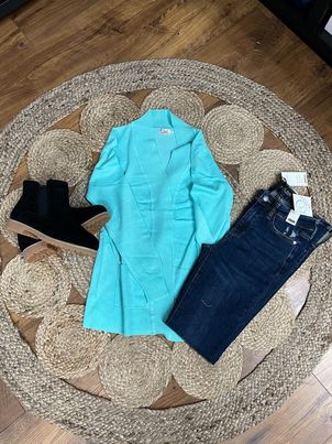 A bright turquoise cardigan laid out on a circular woven mat, surrounded by a pair of black boots and a pair of blue jeans.