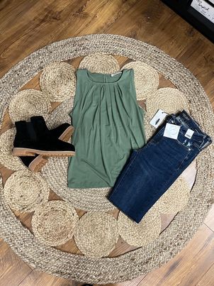 A green pleated tank top displayed on a mannequin, surrounded by accessories including a pair of boots and jeans, presented on a circular straw mat.