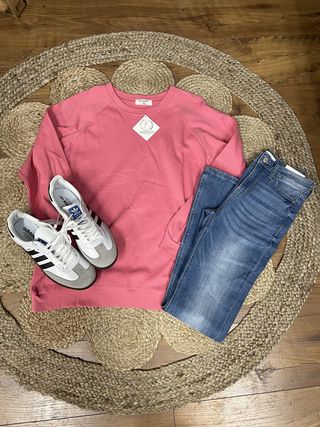 A flat lay of a pink pullover Zenana sweatshirt, blue jeans, and white sneakers with black accents on a woven mat.