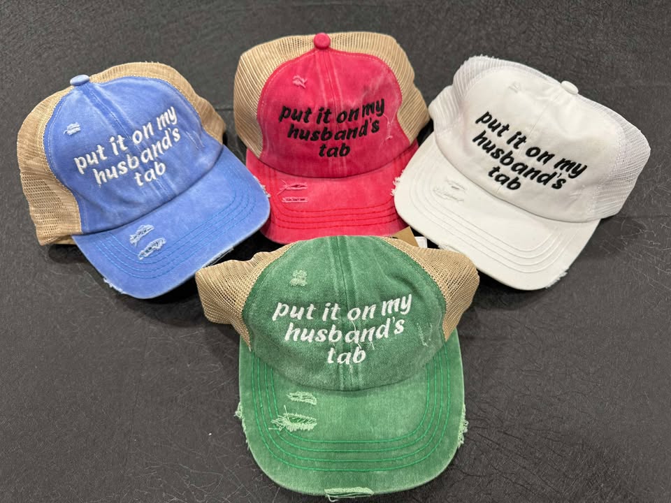 A collection of baseball caps in various colors, each featuring a phrase 'put it on my husband's tab' printed on the front.