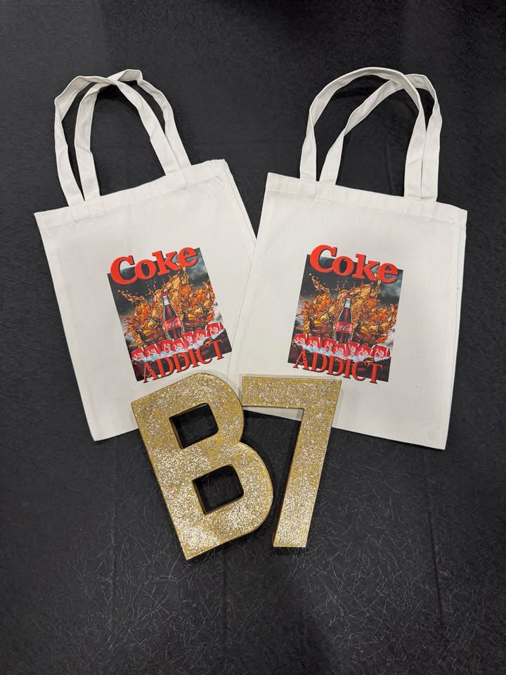 Two white tote bags with 'Coke Addict' text and a graphic design in red and yellow on them, displayed on a dark surface with the letters 'B7' in the foreground.