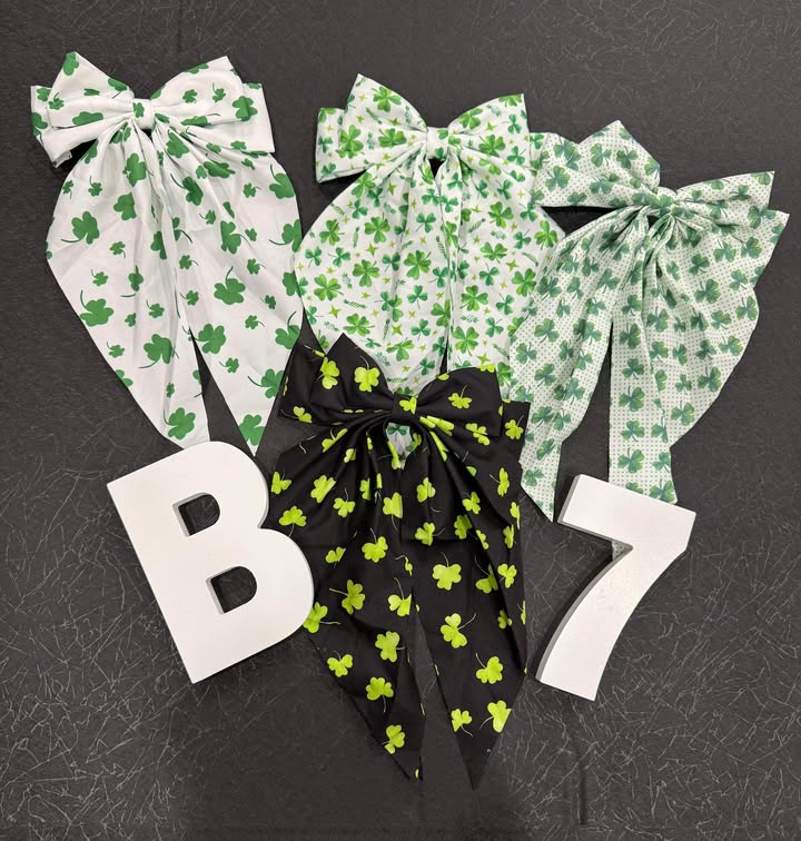 A collection of hair bows with 4 leaf clover patterns in various colors including green and black, displayed on a grey background with letters 'B' and 'L' for branding.