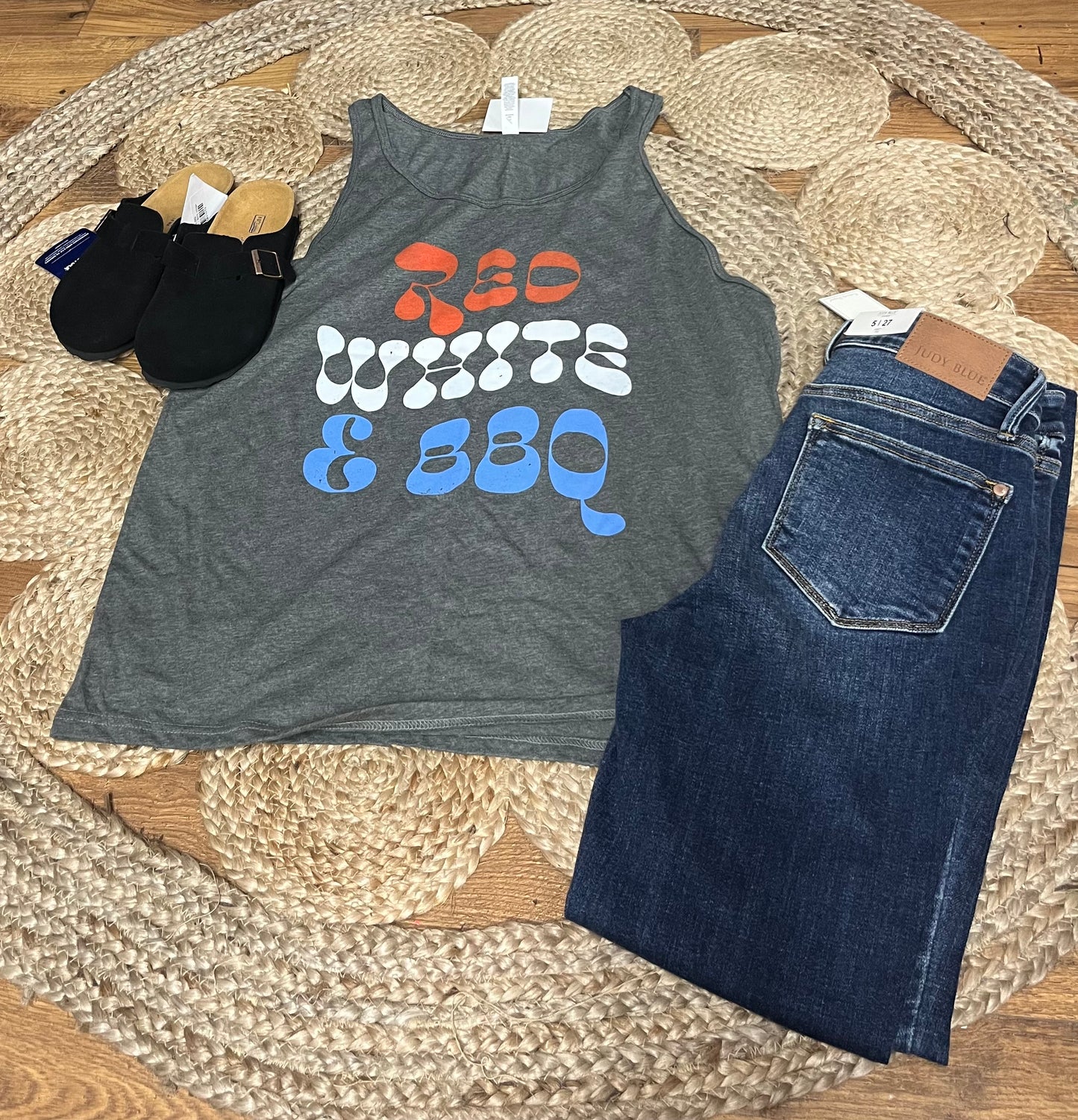 A gray tank top with 'RED, WHITE & BBQ' text in red and white letters laid out flat with a pair of blue jeans and black sandals on a woven mat.