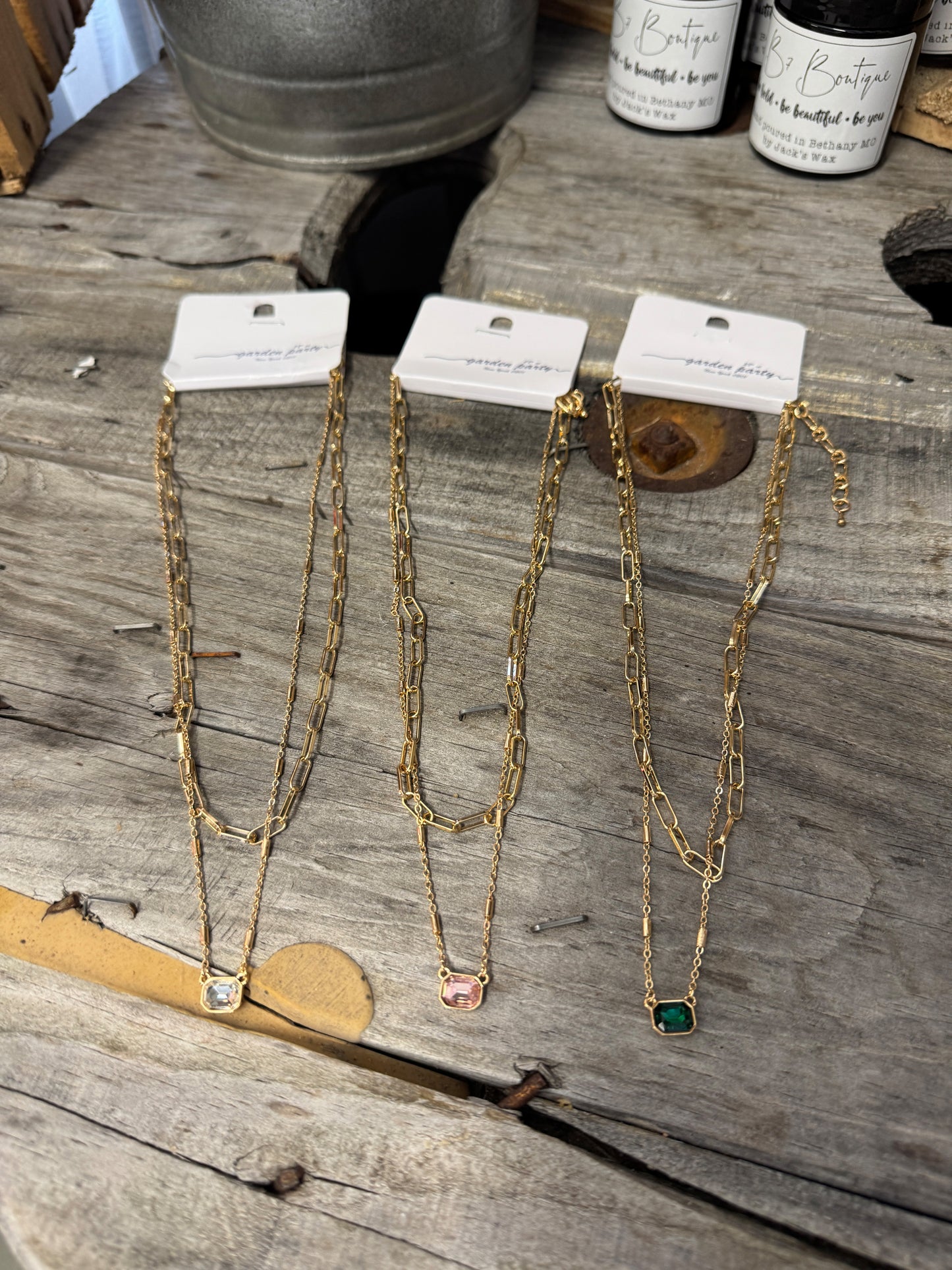 A collection of dainty layered gem necklaces displayed on a wooden surface, with tags indicating price and information.