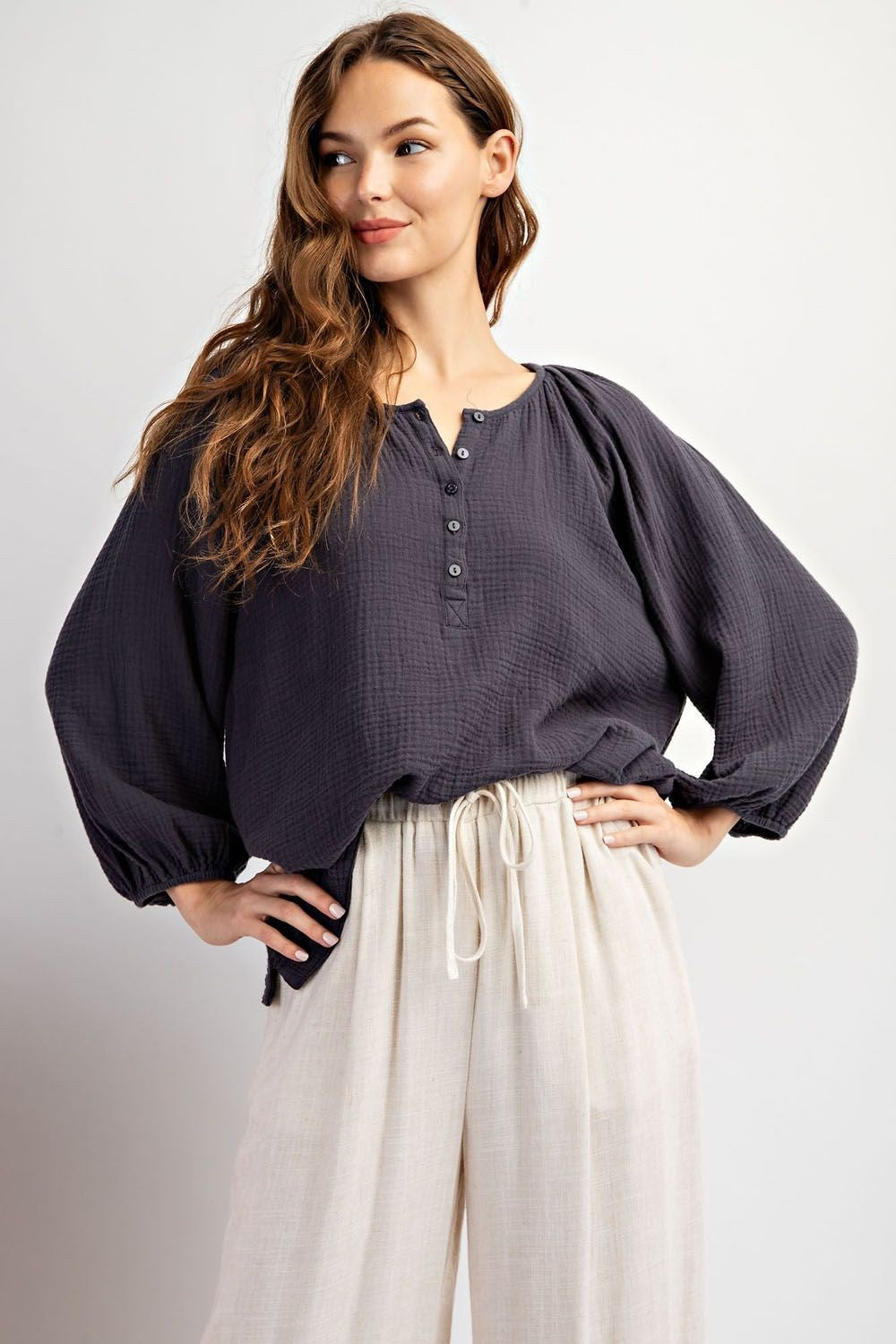 A female model wearing a long sleeve cotton gauze top in a soft washed green color with a round neckline and elastic cuffs, paired with beige trousers.