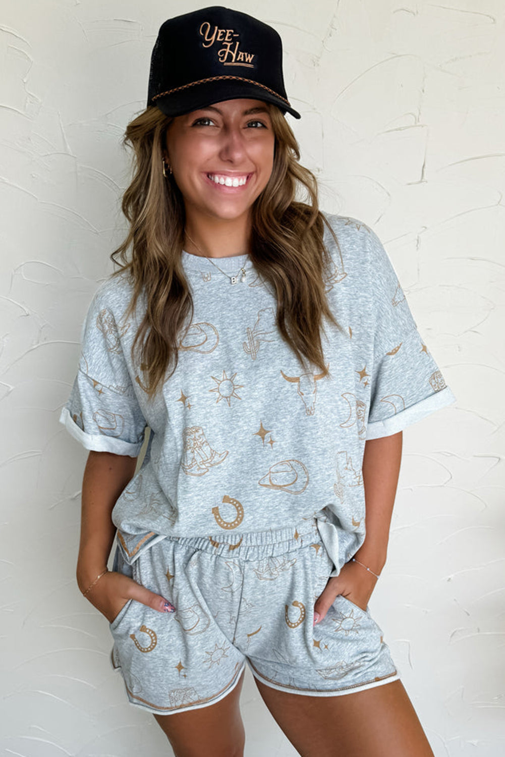 A woman wearing a gray t-shirt with a western fashion print and matching elastic waist shorts, paired with a black cap with text.
