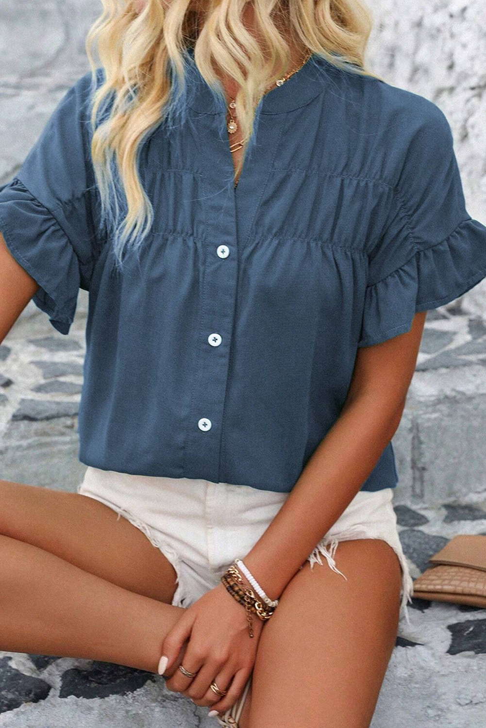 A woman sitting on a rock wearing a blue ruffle ruched short sleeve buttoned shirt with white shorts.