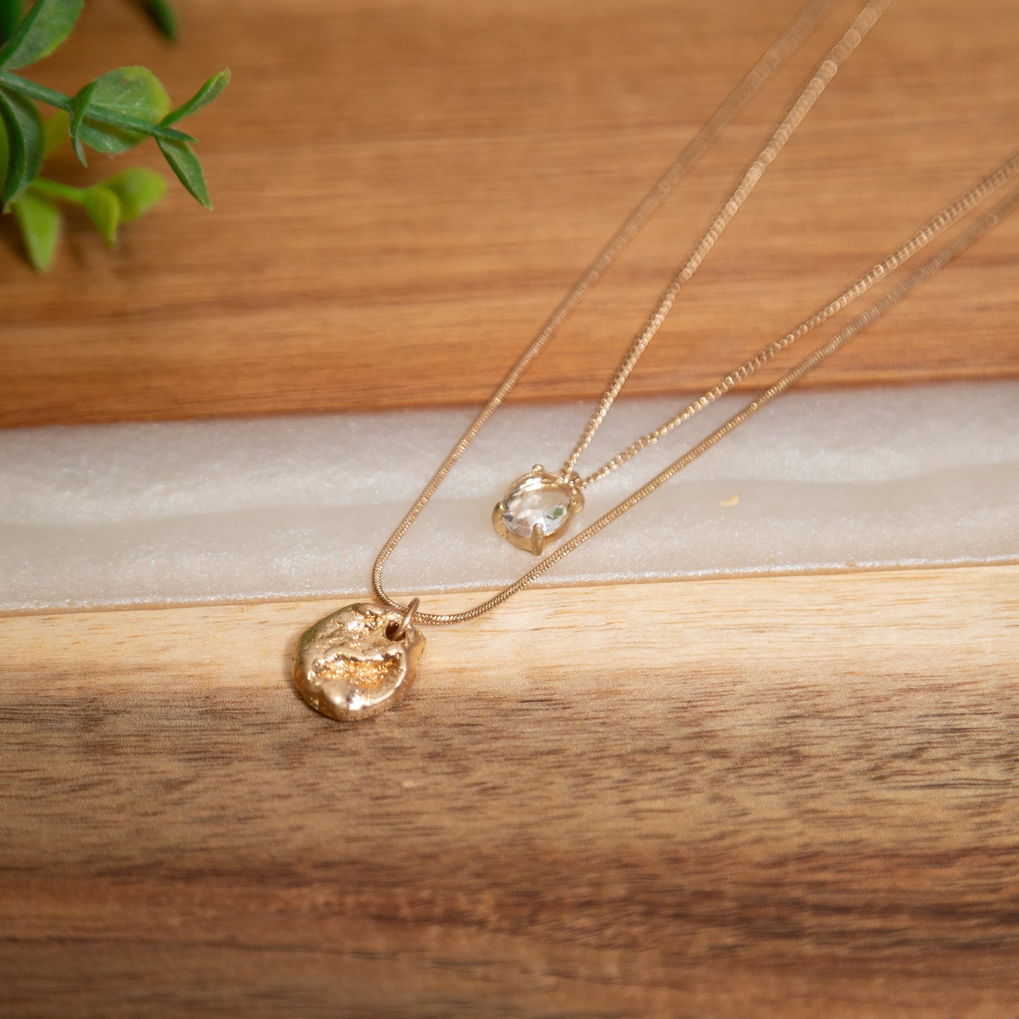 Two gold necklaces on a wooden surface with a plant in the background