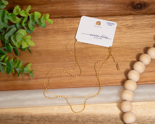 Gold necklace on a wooden surface with a 'garden party' card and a beaded bracelet.
