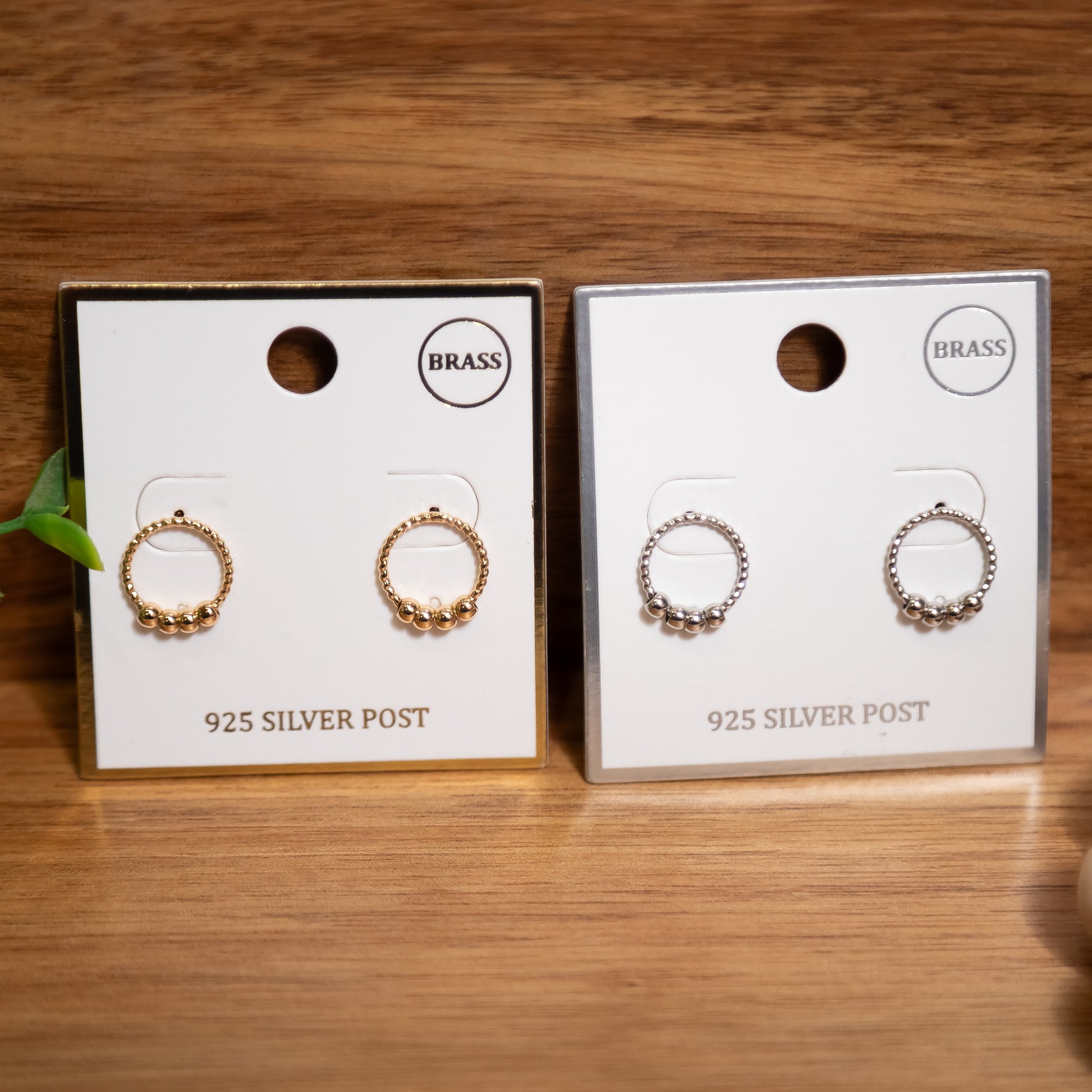 Two pairs of earrings on display cards with 'BRASS' and '925 SILVER POST' text on a wooden surface.