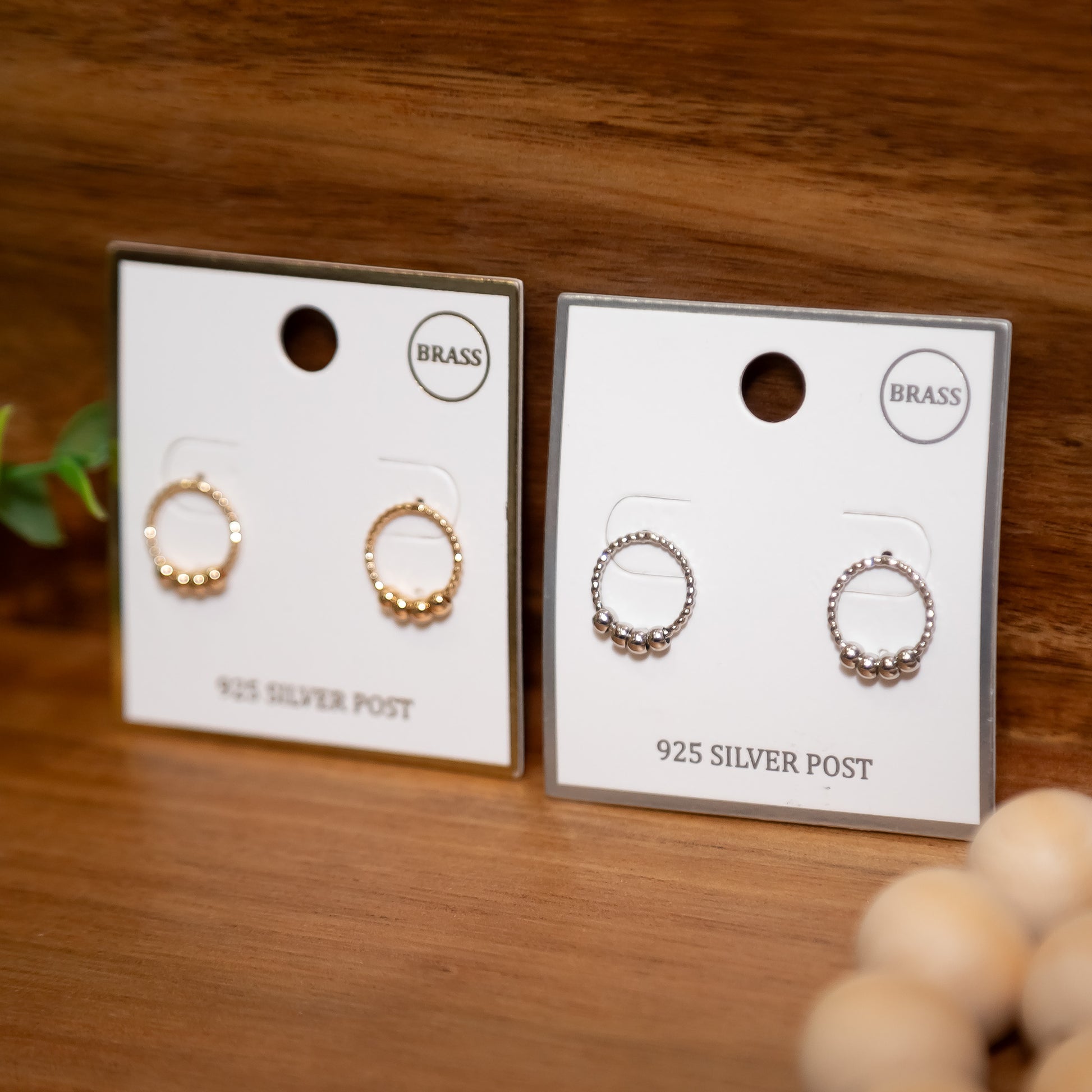 Two pairs of earrings on cards labeled 'Brass' and '925 Silver Post' on a wooden surface.