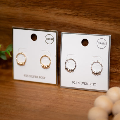 Two pairs of earrings on cards labeled 'Brass' and '925 Silver Post' on a wooden surface.