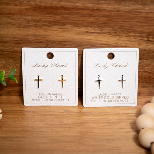 Two pairs of cross-shaped earrings on 'Lucky Charm' cards with gold and white gold finishes on a wooden surface.