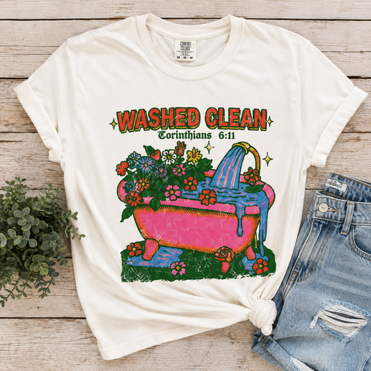 An off-white graphic t-shirt laid flat on a light wood background, styled with a pair of distressed denim shorts and a small green plant. The t-shirt features a colorful retro-style graphic of a pink bathtub filled with flowers and water, with the text 'WASHED CLEAN' and 'Romans 6:11' above it.